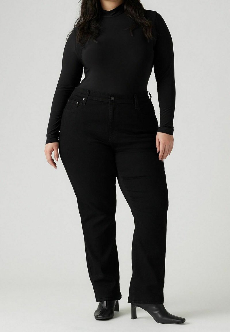 Femme debout portant un haut noir ajusté à manches longues, un jean noir droit et des bottines à talons noires sur fond neutre.