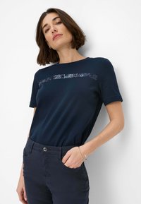 Navyblaues T-Shirt mit kurzen Ärmeln, versehen mit einem horizontalen Paillettenstreifen über der Brust; kombiniert mit dunklen Hosen und einem silbernen Armband.