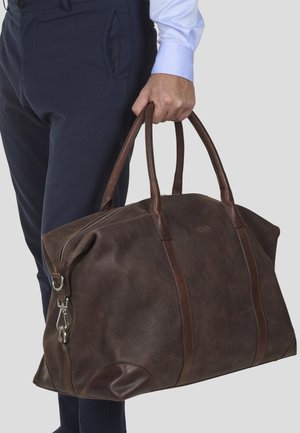 Brown leather duffel bag with dark leather accents, large main compartment, zip closure, and sturdy dual handles. Textured surface.