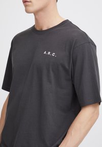 Mørkegrå bomulds t-shirt med korte ærmer, rund halsudskæring og hvid "A.R.C." tekst trykt på venstre brystområde. Casual pasform.
