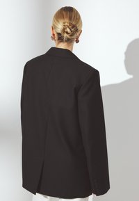 Melna pārmērīga izmēra žakete ar strukturētu dizainu, kurai ir viens aizmugures ventiļs, pielāgotas atloks un pogu aproces. Gluda auduma tekstūra.