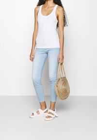 Vit tanktopp med knappdetalj, ljusblå slim fit-jeans, vita sandaletter och en rund strandväska. Slät textur, avslappnad design.