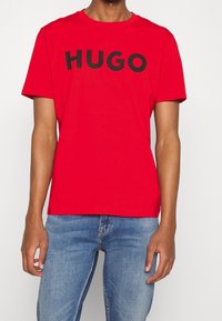 Camiseta de algodón roja con mangas cortas, que presenta un logotipo gráfico en negro "HUGO" en el pecho. Diseño simple de cuello redondo.