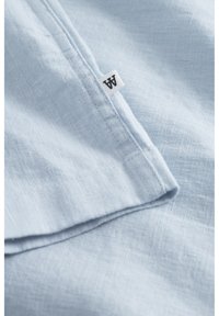 Tissu en lin bleu clair avec une surface texturée, comportant une étiquette blanche avec du texte noir sur le bord. Le tissu est plié et superposé.