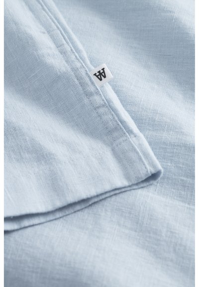 Tissu en lin bleu clair avec une surface texturée, comportant une étiquette blanche avec du texte noir sur le bord. Le tissu est plié et superposé.