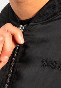 Giacca bomber nera con una texture liscia e lucida. Presenta una chiusura con zip e un logo ricamato "SMILE" vicino alla zip.