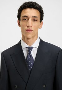 Granatowy garnitur w paski, dwurzędowy z wyłogami, biała koszula i wzorzysty niebieski krawat z małymi białymi kropkami.