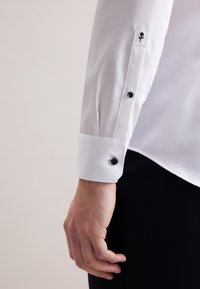 Witte blouse met lange mouwen, manchet met zwarte knopen en een kleine zwarte rozenborduursel nabij de pols, gedragen met zwarte broek.