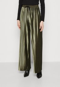 Pantaloni di velluto plissettati verde oliva con vita regolabile, caratterizzati da un design a gamba ampia e una texture liscia. Abbinati a stivali neri con tacco.