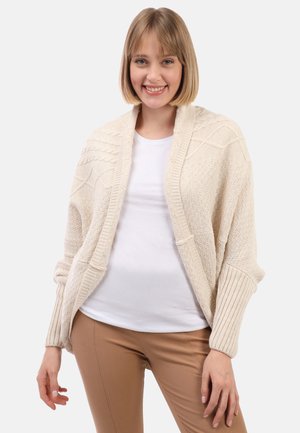 Glimlachende vrouw met kort blond haar, gekleed in een crèmekleurige gebreide cardigan met patroon over een wit T-shirt en lichtbruine broek.