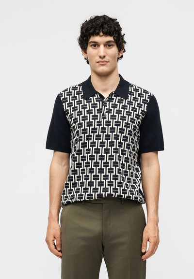Man wearing black and white geometric patterned short-sleeve polo shirt and olive-green pants, standing against a plain background.
