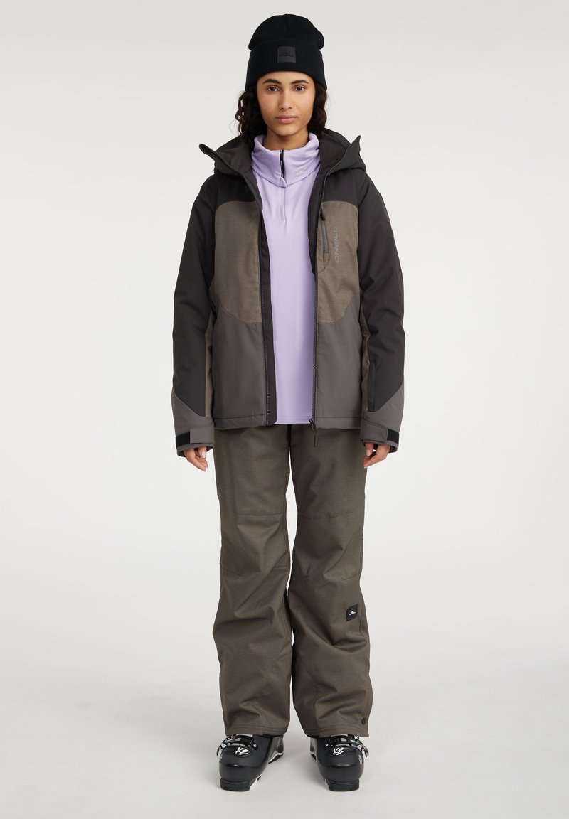 Skijacke in Schwarz und Braun mit weicher Textur, Reißverschluss und Kapuze. Eine lockere olivegrüne Skihose und schwarze Stiefel.