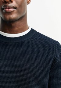 Marineblau gestrickter Pullover mit einem strukturierten Muster, geripptem Kragen und weißem Innenfutter. Nahaufnahme mit Fokus auf dem Halsbereich und der oberen Brustpartie.