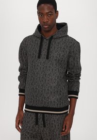 Sudadera con capucha negra que presenta un patrón texturizado con una marca en blanco, puños y dobladillo acanalados con una franja contrastante. Diseño de corte holgado.