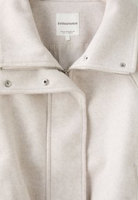 Gros plan sur le col d'une veste en laine beige montrant une fermeture éclair métallique partiellement fermée et des boutons-pression, avec une étiquette de marque en tissu à l'intérieur.