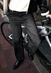 Jeans neri a gamba larga con texture liscia, indossati con stivali neri, posizionati accanto a una bicicletta bianca su una superficie in mattoni.