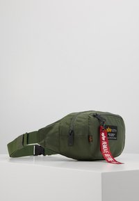 Marsupio in nylon verde con superficie testurizzata, strap regolabile, chiusura con zip e un'etichetta in tessuto rossa con testo. Presenta un piccolo logo.