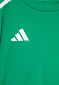 Camiseta atlética verde con cuello redondo. Presenta un gran logo de Adidas en blanco, hecho de un material suave, y un acento blanco en contraste en el hombro.