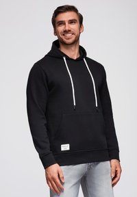 Černá mikina s kapucí vyrobená z bavlněné směsi, která má klokaní kapsu, bílé šňůrky a malou značkovou nálepku na přední straně. Hladká textura.