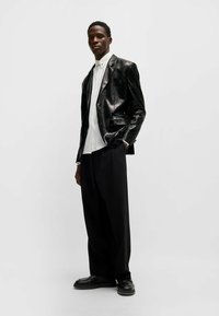 Schwarzer Pailletten-Blazer über einem weißen Hemd, kombiniert mit lockeren schwarzen Hosen und schwarzen Schuhen. Der einfarbige Hintergrund hebt das Outfit hervor.