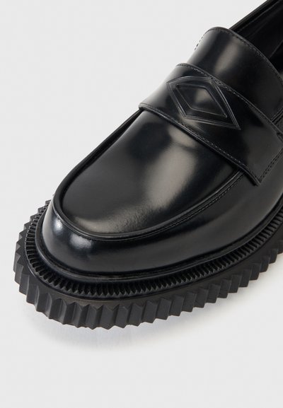 Μαύρο λουστρινένιο μοκασίνι με ανάγλυφο διακοσμητικό σε σχήμα διαμαντιού και παχιά σόλα από καουτσούκ με ραβδώσεις.