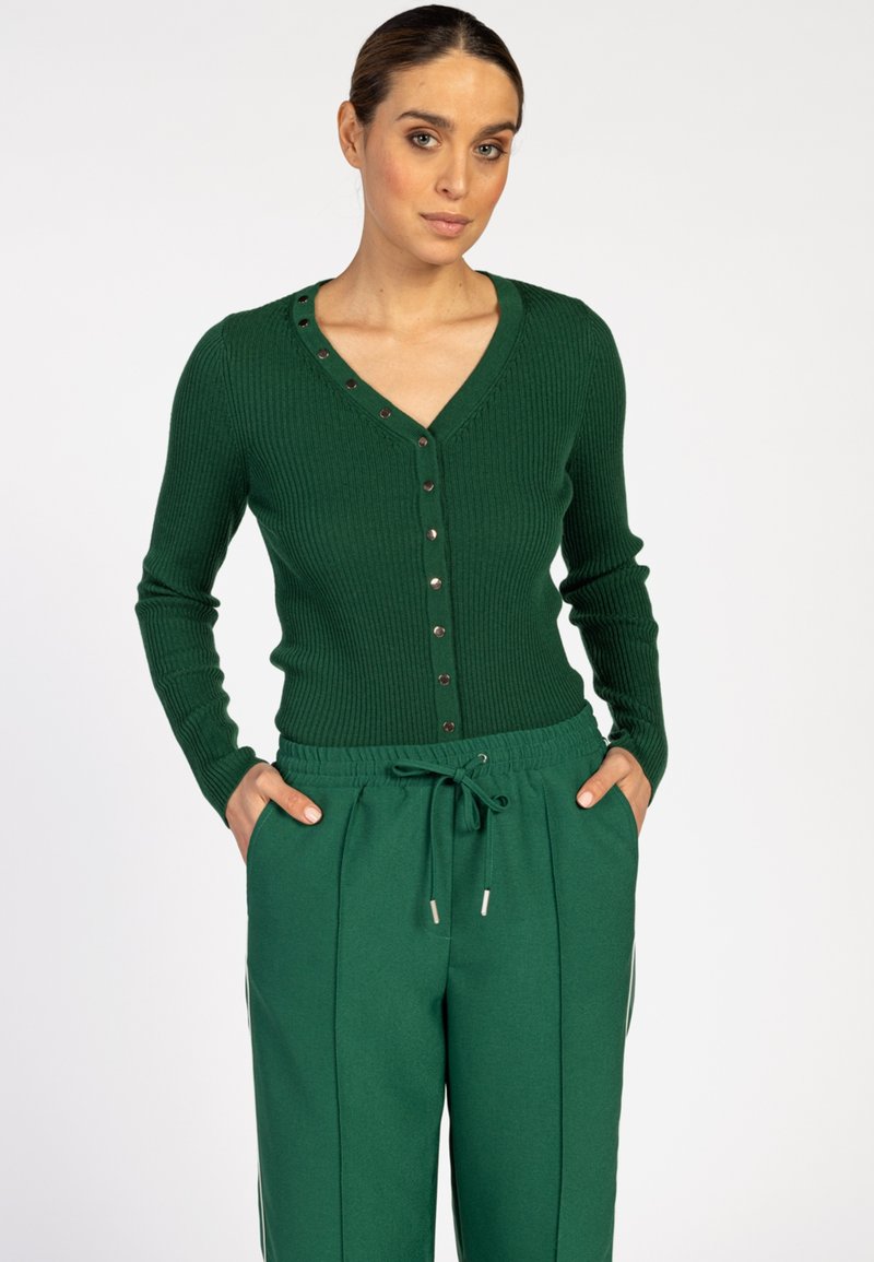 Vrouw in een donkergroene ribgebreide cardigan met knopen en bijpassende groene broek met koord, staand met handen in de zakken tegen een witte achtergrond.