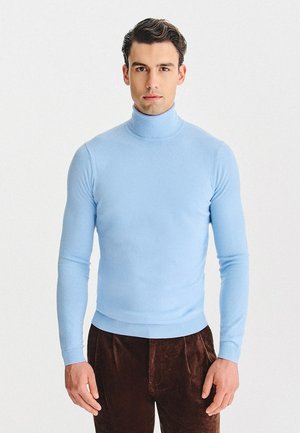 Jasnoniebieski sweter z golfem wykonany z miękkiego materiału, z długimi rękawami i dopasowanym krojem, zestawiony z ciemnobrązowymi spodniami z kordurowej tkaniny.