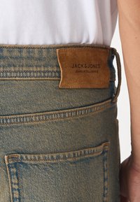 Lyseblå denimjeans har en brun semsket lærpatch på livet, preget med "JACK & JONES JEANS INTELLIGENCE" i mørk skrift.