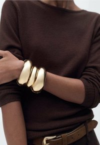 Bracelet manchette métallique ton or avec trois sections lisses et courbées. Porté au poignet avec un pull en maille marron et une ceinture.