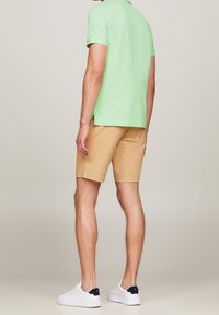 Homme portant un polo vert clair, un short beige et des baskets blanches, debout de dos face à l'appareil photo sur un fond uni.