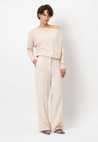Beige off-shoulder trui met decoratieve knopen, gecombineerd met crèmewitte broek met wijde pijpen. Schoenen hebben een zwart en roze zebra-patroon.