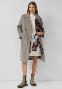 Cappotto doppiopetto in lana grigia, abito crema, sciarpa a quadri legata in vita, pochette lucida color bordeaux e stivaletti marroni con suole nere.