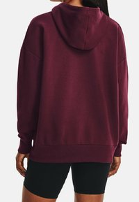 Persona che indossa un felpa larga color bordeaux e pantaloni shorts neri attillati, mostrata di spalle su uno sfondo neutro.