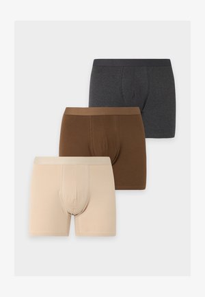 Homme à la peau foncée et aux cheveux courts et bouclés, debout pieds nus portant un boxer beige sur un fond gris uni.