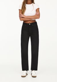 Jeans negros de talle alto con diseño de pierna recta, estilo de cinco bolsillos y cierre de botón plateado, combinados con zapatillas blancas.