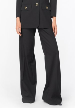 Kvinde iført sorte bukser med vide ben og matchende sort blazer med guldknapper, stående mod en hvid baggrund.