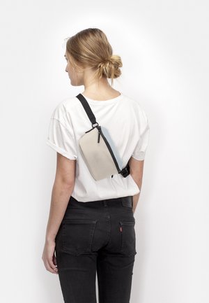 Young woman with blonde hair in a bun wears a white t-shirt and black jeans, carrying a beige crossbody bag with a black strap on her back.