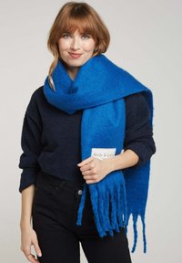 Écharpe en laine bleue avec des bords à franges, texture douce, portée autour du cou. L'étiquette indique "Andy & Lucy." Portée avec un haut sombre et un pantalon noir.