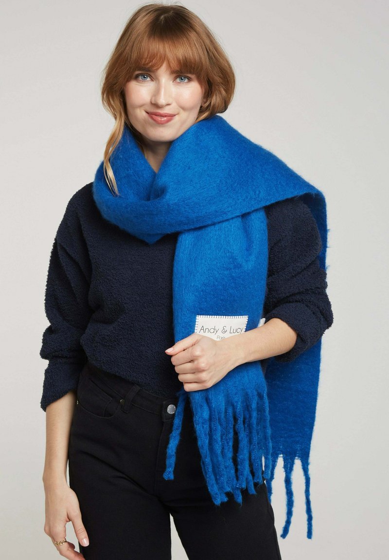 Écharpe en laine bleue avec des bords à franges, texture douce, portée autour du cou. L'étiquette indique "Andy & Lucy." Portée avec un haut sombre et un pantalon noir.