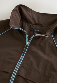 Veste marron avec bordure bleue, fermeture éclair argentée partiellement ouverte et une étiquette noire "NEXT" à l'intérieur du col.
