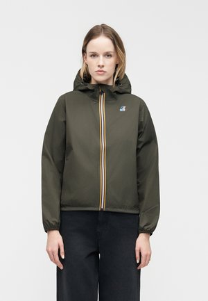 Woman wearing a green hooded jacket with a striped zipper and logo on the chest, standing against a plain gray background.