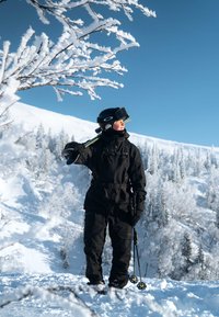 Completo da sci in tessuto nero con cerniere e tasche, indossato con casco nero e occhiali da sci. Circondato da alberi e montagne innevate.
