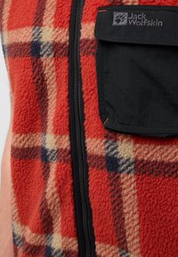 Rote Fleece-Weste mit einem Karo-Muster in Creme und Marineblau, ausgestattet mit einem schwarzen Reißverschluss und einer Tasche. Weicher Stoff und warmes Design, ideal für das Layering.