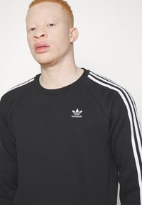 Sweatshirt noir avec le logo Adidas blanc et trois bandes blanches sur chaque épaule. Fabriqué en tissu doux avec un col ras du cou et des manches raglan.