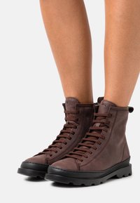 Hnědé kožené šněrovací boty s elegantním designem, vysokým kotníkem a černou gumovou podrážkou, která má texturovaný dezén pro lepší přilnavost.