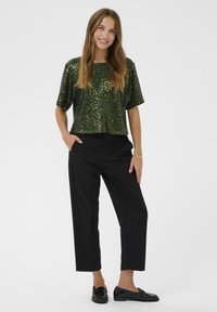 Vrouw die glimlacht, gekleed in een kortmouwige donkergroene sequintop, zwarte cropped broek en zwarte glanzende instappers, staande met handen in de zakken.