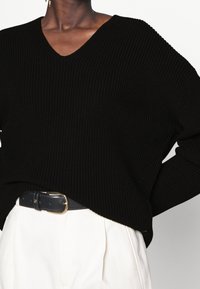 Svart ribbad stickad tröja med V-ringning, med en lös passform och ärmar, ihop med högmidjade ljusa byxor och ett svart bälte.