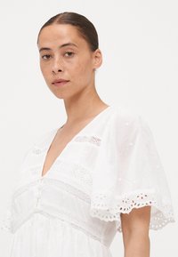 Il vestito bianco presenta un profondo scollo a V, maniche corte a campana, strisce orizzontali ricamate e un bordo decorativo a occhietti lungo le maniche.