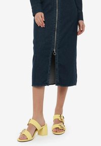 Abito midi in denim blu scuro con zip frontale e spacco. Indossato con sandali platform gialli con grandi fibbie.