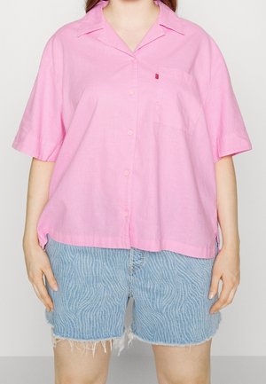 Person iført en løs lyserød kortærmet skjorte med knapper og lyseblå mønstrede denimshorts med frynser i kanten.